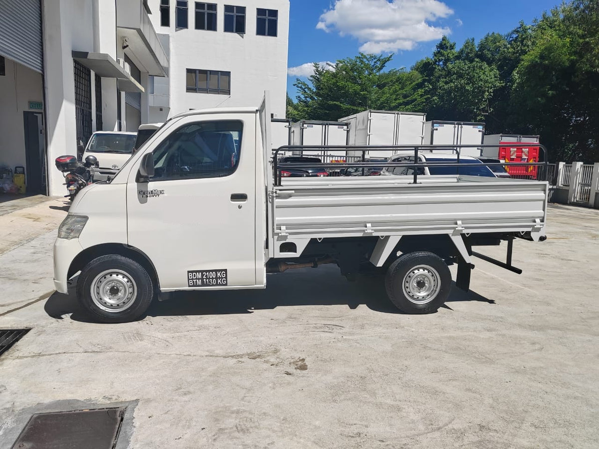 A white Daihatsu Gran Max compact pickup truck with a silver metal cargo bed and black tires, parked outdoors on a concrete lot next to a white building under a blue sky with clouds.
