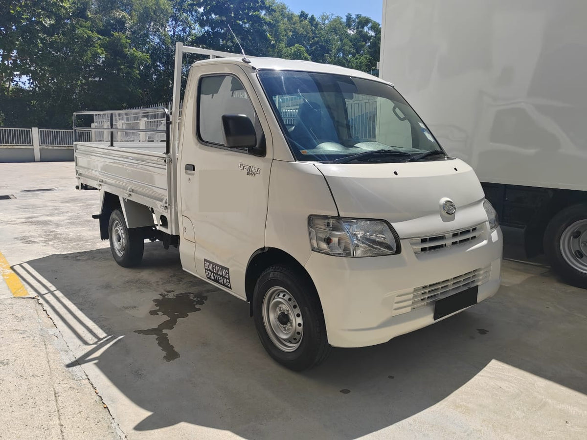 A white Daihatsu Gran max single-cab utility truck parked on a concrete lot next to a large white truck, with trees in the background.