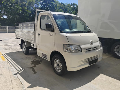A white Daihatsu Gran max single-cab utility truck parked on a concrete lot next to a large white truck, with trees in the background.