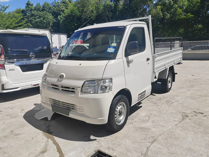 A white Daihatsu Gran Max steel cargo truck parked outdoors on a concrete lot, with other vehicles and green trees visible in the background.