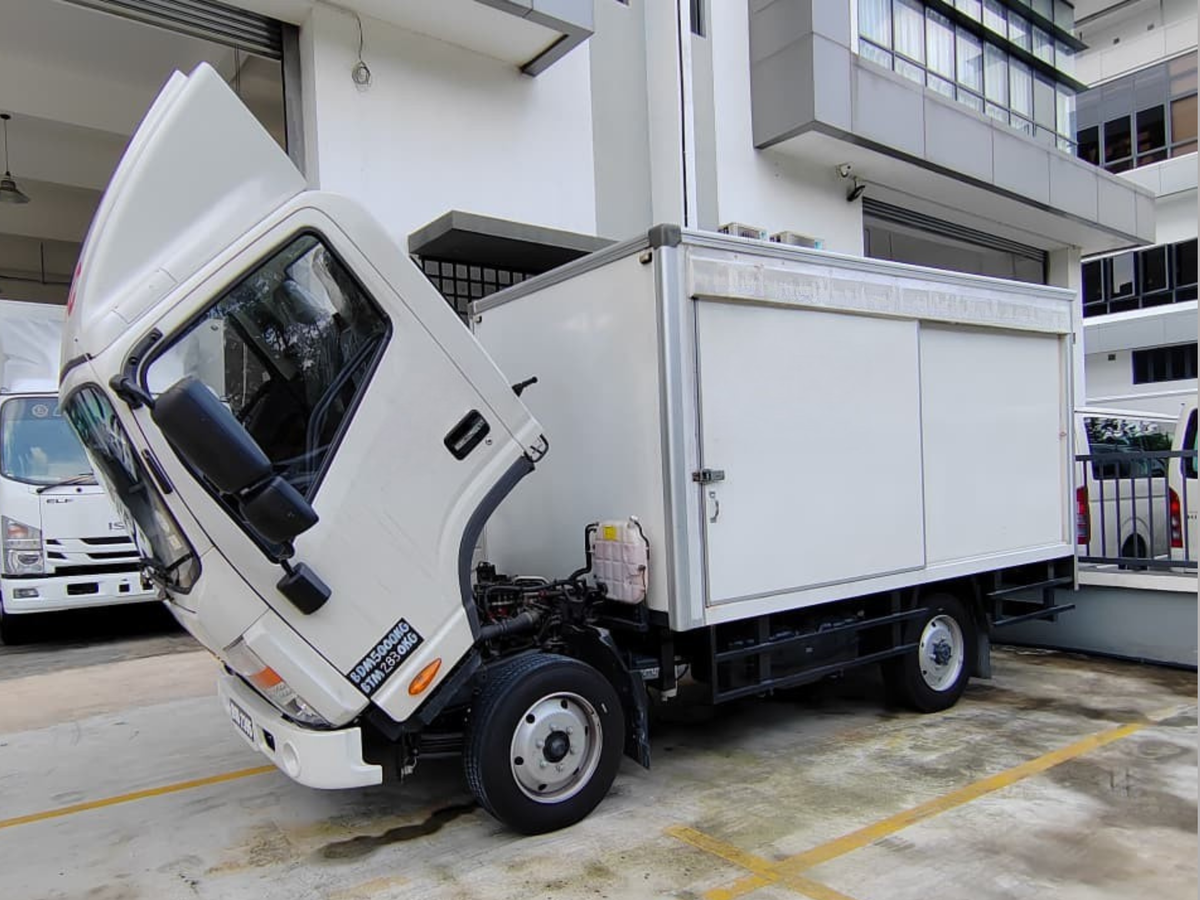 A white JAC N50 box truck parked in a lot outside a building, with the driver's side door open and the front of the cab tilted upward. The truck has a white cargo body, black wheels, and a small refrigerant tank visible beneath the cab.