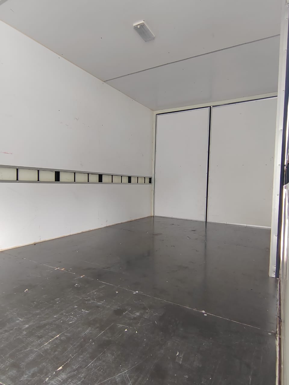 An empty white van or trailer interior with a dark gray floor, white walls, a sliding door, and a ceiling light.