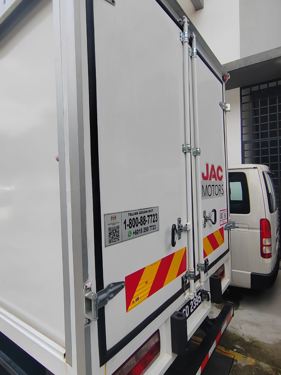 White JAC Motors box truck rear cargo door with silver hinges and red-and-yellow reflective striping, photographed in a warehouse or garage setting next to another white vehicle.