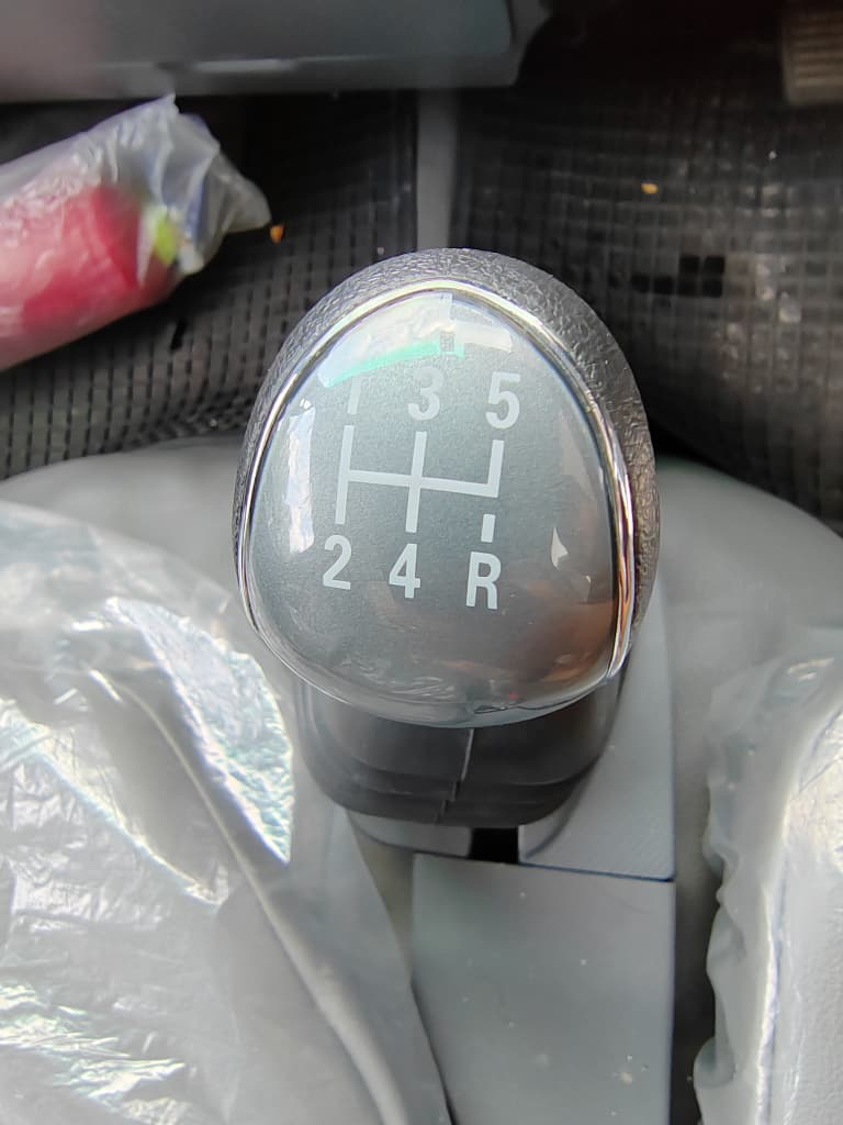 A 5-speed manual gear shift selector knob inside a car, showing white shift markings (R, 2, 3, 4, 5) on a gray face with a chrome bezel.