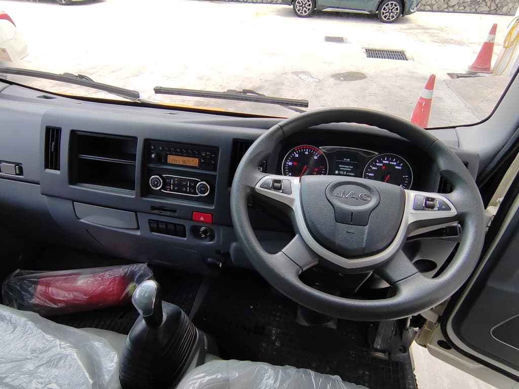 A JAC vehicle interior dashboard and steering wheel view, with the driver's seat door open, showing the center console, instrument cluster, gear shift, and a paved outdoor parking area outside.