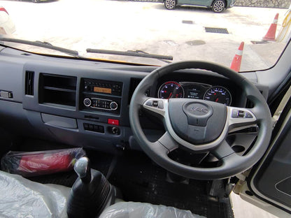 A JAC vehicle interior dashboard and steering wheel view, with the driver's seat door open, showing the center console, instrument cluster, gear shift, and a paved outdoor parking area outside.
