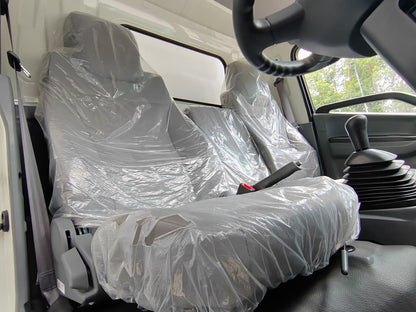 A vehicle interior with two front seats covered in clear plastic protective wrapping, along with a steering wheel, gear shift knob, and side door panel visible.